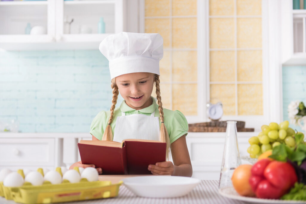 Young girl wears a chef hat while looking through a cookbook.